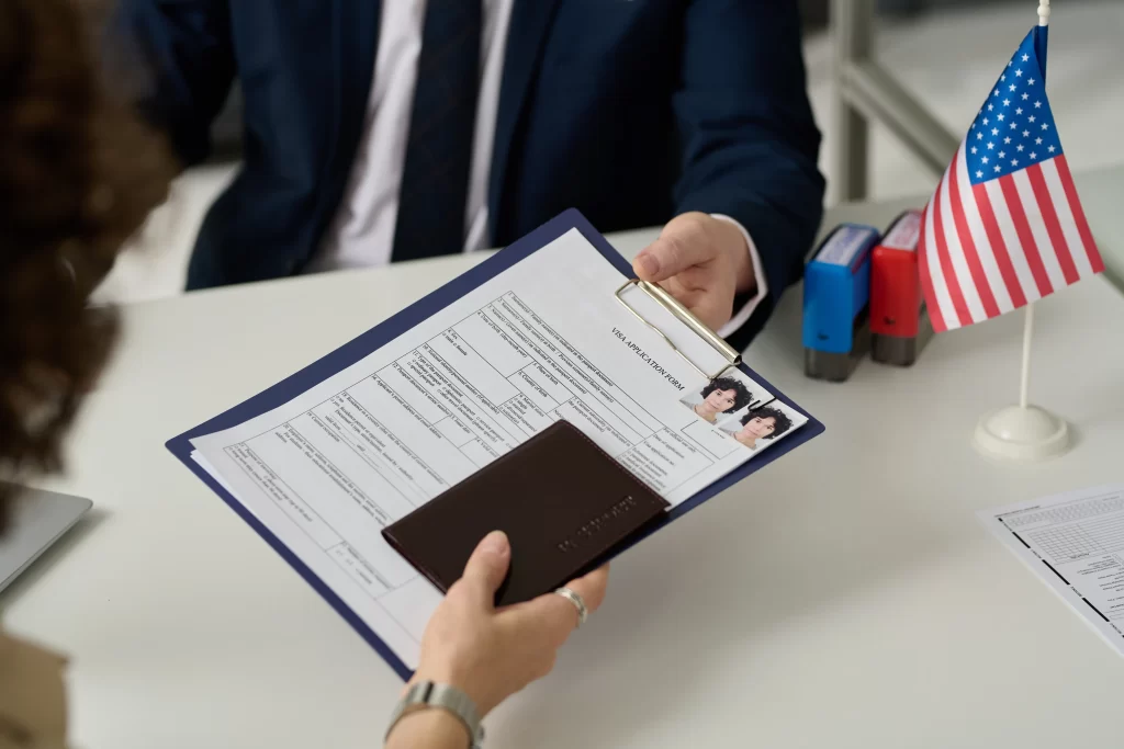 Mujer entregando tablero con documentos, pasaporte y fotos a hombre en traje y corbata quien tiene una bandera de Estados Unidos en su escritorio