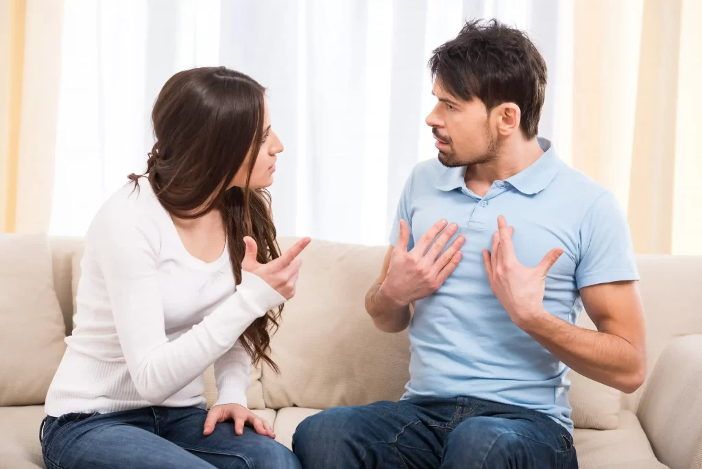 Couple consisting of a man and a woman arguing sitting on a coach. The woman points to the man, and the man points to himself