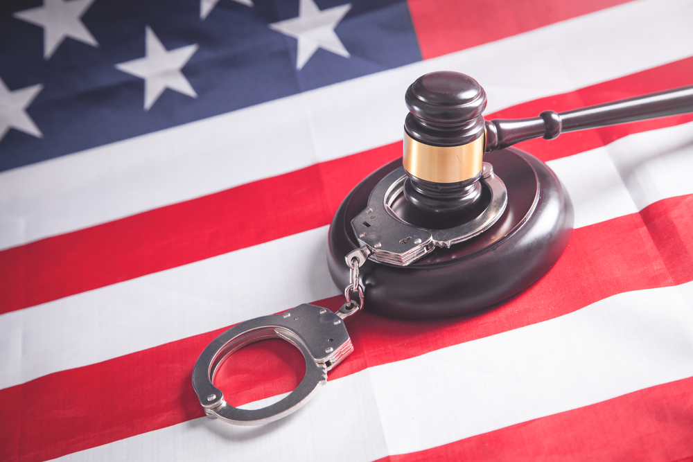 A gavel on a table with an American flag and handcuffs.