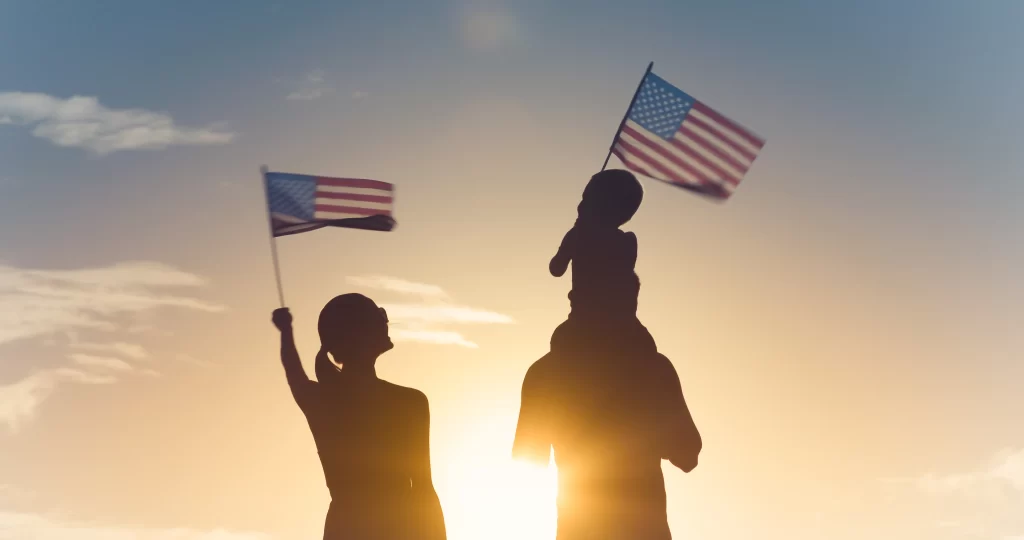 Silhouette of a woman, a man and a child waving US flags with sunset in the background