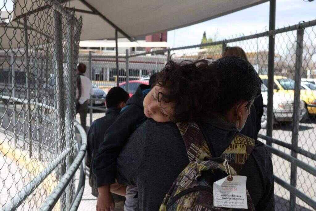 Woman carrying her son as she walks in line behind other migrants along a fenced path at the US-Mexico border